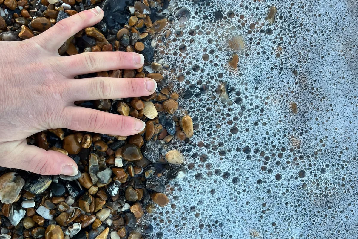 A hand resting on wet shingle at the edge of the sea, foam breaking around it